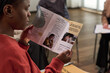 © pressmaster - Young adult Black woman reading informational brochure about group therapy while sitting in support group session, holding pamphlet with mental health resources, other people visible in background