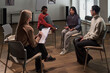 © pressmaster - Group of adults of diverse ethnicities sitting in circle discussing during support group session, Caucasian woman holding clipboard listening attentively