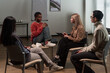 © pressmaster - Group of adults and one Black gen Z girl sitting in circle engaging in discussion during support group meeting, female facilitator holding clipboard listening attentively