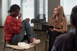 © pressmaster - Black teenager sitting with knees up, holding face in hand, showing distress while Caucasian middle aged woman therapist holding clipboard, listening attentively during counseling session