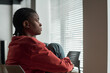 © pressmaster - Black woman sitting by window gazing outside with knees drawn up, wearing casual clothing, natural light streaming through blinds, depressed expression visible on face