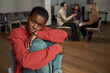 © pressmaster - Portrait of Black young adult woman sitting with knees up looking into camera while group of diverse adults having discussion in background, support meeting in modern room