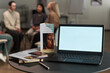 © pressmaster - Open laptop with blank screen standing on table near brochures and notebook, background showing group of multiethnic people sitting in circle talking during therapy session
