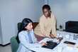 © pressmaster - Hispanic young adult woman sitting at desk using laptop while Black young adult woman standing beside her pointing at document, both collaborating on business project in office