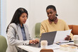 © pressmaster - Hispanic young adult woman and Black young adult woman collaborating at table, working on laptop and reviewing documents, both focused on project, coffee cups and papers visible