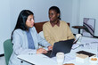© pressmaster - Hispanic young adult woman working on laptop while Black young adult woman discussing documents at office desk, both focused on business collaboration