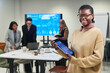 © pressmaster - Young adult Black woman smiling and holding digital tablet with data charts, standing in modern office while multiethnic colleagues working and discussing analytics in background