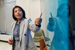 © pressmaster - Young adult Hispanic woman explaining concept while standing at glass board, holding marker and gesturing toward diagram, business setting with office chair in background
