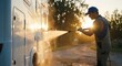© Gayan - Adult white man in a blue cap power washing a white camper van outdoors during golden hour sunset, professional RV cleaning and maintenance service concept, bright sun flare lighting.