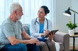 © DragonImages - Caucasian senior man sitting on sofa talking with Hispanic female doctor holding digital tablet, medical professional listening attentively and taking notes during consultation