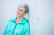 © Westend61 - Smiling sporty mature woman with earbuds leaning against concrete wall