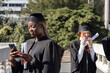 © wavebreak3 - Diverse female graduates in gowns and caps standing on bridge holding diplomas and checking phone