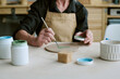 © AnnaStills - Senior woman painting ceramic plate with brush, holding paint container in hand, sitting at table with jars of paint and sponge, focusing on creative pottery work