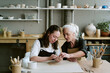 © AnnaStills - Caucasian young adult woman and Caucasian senior woman sitting at table shaping clay together in pottery studio, both focused on creative process