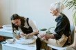 © AnnaStills - Young adult Caucasian woman shaping clay on pottery wheel while senior Caucasian woman observing and guiding her, both sitting in ceramics studio with pottery tools