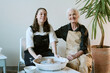 © AnnaStills - Caucasian young adult woman shaping clay on pottery wheel while senior Caucasian woman sitting beside her smiling, both wearing aprons, indoor creative activity, plant in background