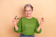 © deagreez - Confident mature woman with short grey hair in green sweater smiling while holding two pairs of glasses against a beige background for lifestyle and fashion marketing