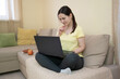 © Yuliia - Young woman sitting cross-legged on a sofa at home, using a laptop. Smiling woman working remotely or browsing the web. Cozy interior, casual lifestyle, and digital connectivity.