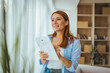 © Dragana Gordic - Smiling Woman Using Tablet At Home Near Window, Remote Work And Relaxed Living Room Lifestyle