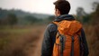 © Bussakon - A solitary backpacker with a vibrant orange backpack journeys along a dusty trail through a muted natural landscape viewed from behind