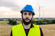 © Xavier Lorenzo - Young man wearing hard hat and safety vest standing in front of wind turbines, representing sustainable innovation. Renewable energy and industrial workforce concept