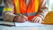 © miss irine - Worker in safety vest and hard hat signs contract on desk. Person fills out paperwork, signs document for work or permit. Blue collar job approval for shift.