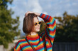© SHOTPRIME STUDIO - Woman portrait in a colorful rainbow sweater and yellow sunglasses smiling outdoors, casual fashion look with short hair and hand on head, bright natural light and relaxed mood.