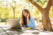 © maxbelchenko - A smiling woman with a laptop sits at a wooden table in a sunny park. A female freelancer works on a laptop and phone outdoors. Concept: education, remote work. Lifestyle.