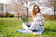 © maxbelchenko - Joyful woman sits with yellow notebook at table in blooming garden. Young woman works on laptop, holding notebook, on spring day. Concept of freelancing, relaxation, and weekends.