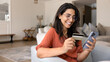 © fizkes - Happy Brazilian woman sits on armchair with smartphone, holding debit card, booking travel or accommodation, shopping through electronic commerce website or app, renewing digital services subscription