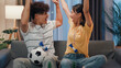 © tirachard - Asian new marriage couple sit on sofa excited cheering watching football sport game on television in living room at home. Young couple sport fans hold football watching online soccer on TV.