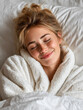 © SvoP - Young woman enjoying a cozy moment wrapped in a soft bathrobe with eyes closed and a peaceful smile while resting in bed under white blankets