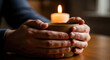 © hilwanstock - Close-up of hands holding a lit candle in a dimly lit room with warm tones.