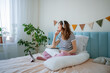 © wifesun - Pregnant woman comfortably sitting on her bed, wearing headphones and using a tablet to participate in an online lecture about childbirth, diligently taking notes in a notebook for birth education