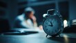 © dinda - Alarm Clock on Desk with Tired Person Working Late at Night.