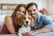 © oksa_studio - Cheerful couple and their pet beagle dog sitting on a sofa, embracing family life and happiness together indoors