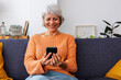 © Xavier Lorenzo - Happy senior woman enjoying funny social media content on mobile phone app while relaxing on cozy sofa at modern apartment. Technology lifestyle concept