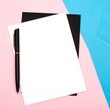© Iwel - A white sheet of paper sits atop a black notepad, with a pen resting on its left side. The background features a pastel pink and blue