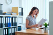 © pressmaster - Caucasian young adult woman working in modern office, reading documents at desk with computer and plant, shelves with organized binders and folders in background, focused expression