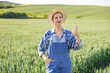© sofiko14 - A smiling woman wearing overalls and a straw hat gives a thumbs-up gesture while standing in a lush green field of crops under a clear sky