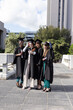 © wavebreak3 - Diverse female graduates posing on campus terrace in black gowns caps holding smartphone, scrolls
