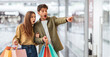 © Prostock-studio - Two people stand in a shopping mall holding colorful bags. They look surprised and excited as they point to something in front of them while standing close together.