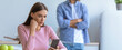 © Prostock-studio - A young woman focuses on her smartphone while sitting at a table in a kitchen. A man stands in the background with his arms crossed, looking upset.