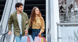 © Prostock-studio - A man and woman are holding colorful shopping bags while walking down an escalator in a mall. They are smiling and enjoying their shopping trip together in a busy area.