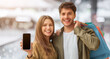 © Prostock-studio - A couple stands inside a shopping mall smiling. The man holds a smartphone in front of him while the woman stands beside him. They are both happy and holding shopping bags.