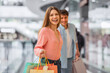 © Prostock-studio - A woman smiles while holding colorful shopping bags as she walks through a busy mall with a man nearby. They appear happy and engaged in a fun activity on a weekend.