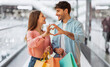 © Prostock-studio - Man and woman smile at each other in a mall. They hold shopping bags. They form a heart shape with their hands. The atmosphere is lively and fun as people walk by.