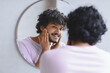 © Prostock-studio - A man stands in front of a round mirror while checking his skin. He gently touches his face and smiles. Sunlight illuminates the room, creating a bright atmosphere for grooming.