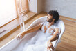 © Prostock-studio - A man sits in a bathtub filled with bubbles. Sunlight comes through the window, and the bathroom has a modern design with wooden floors. He appears to enjoy a moment of relaxation.