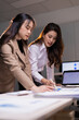 © Tj - Two businesswomen are actively collaborating, analyzing business charts and financial data on documents and a laptop, discussing strategies and financial reports in a professional office environment